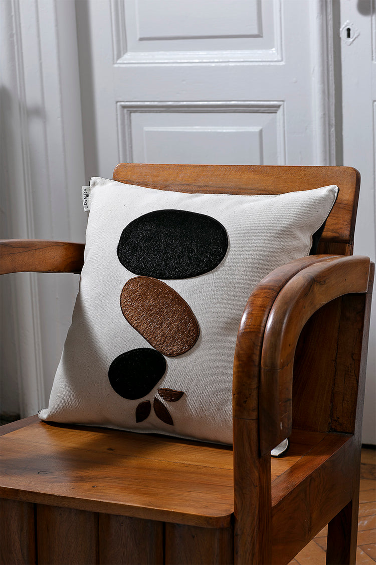 Minimalist bohemian decorative pillow with black and brown abstract pebble design, placed on a rustic wooden chair. Modern boho style accent pillow, perfect for living room or bedroom decor.